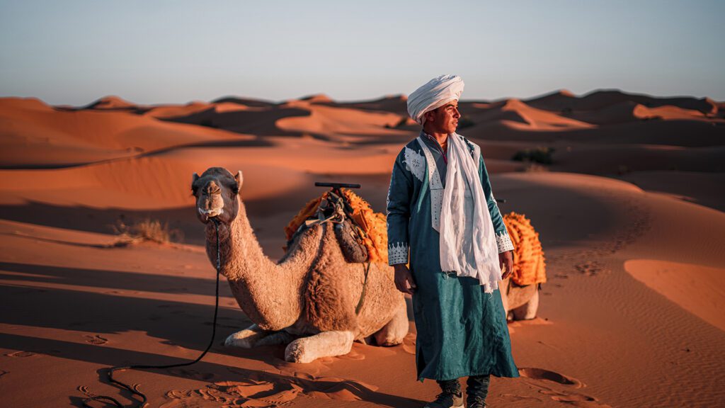 A man in traditional Berber clothing walks in the Sahara Desert in Morocco. He is wearing a white turban and a robe that is green/blue in shade with white decoration. Two camels sit on the sand behind him. The man looks to his left, toward the sun. The long shadows extending in the opposite direction suggest that it is dusk. In the distance, behind the man and the camels, the burnt orange dunes roll off toward the horizon.