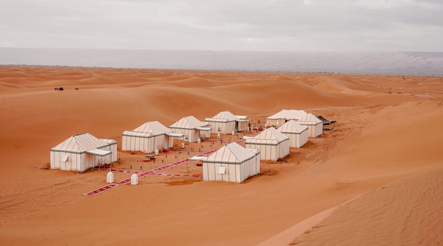 An elevated view of Desert Luxury Camp's Erg Chigaga luxury desert camp. Two rows of four luxury tents face one another across the sand. A central line of red patterned rugs runs between the two rows, leading toward another large tent pitched at one end. Rugs cross the central pathway, leading to the entrances of each tent. The rolling golden dunes of Morocco's Sahara Desert surround the campsite.