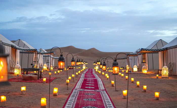A ground-level view from inside the Main Camp at Desert Luxury Camp's Erg Chigaga site in Morocco. It's dusk, so the sky is darkening. The towering dunes in the middle distance are also a darker shade of gold. In the foreground, a line of red patterned rugs stretches toward the dunes, but stops short. On either side of this rug pathway are large luxury tents. They are white in colour, with dark stripes and edging. Many lanterns, some suspended on poles lining the rug pathway, others placed on the sand, glow with golden light, creating a comfortable, cosy atmosphere.