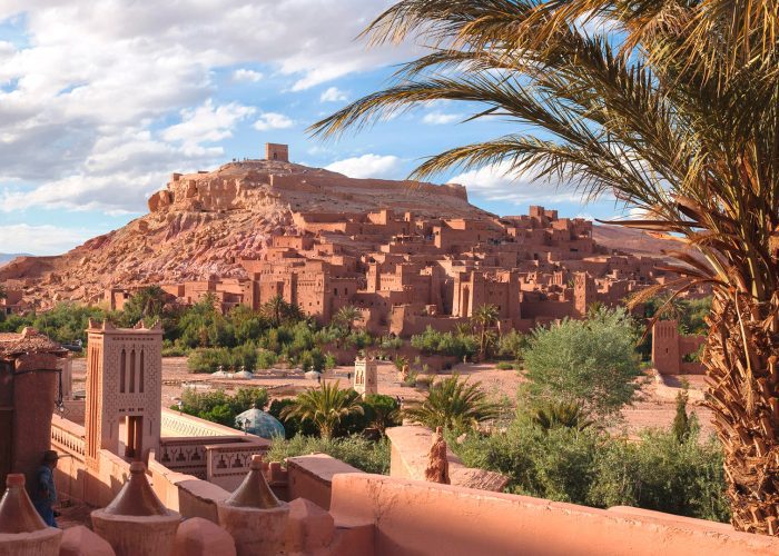 The historic ighrem, or ksar, of Aït Benhaddou in Morocco viewed from a distance. A nearby palm tree frames the view of the small fortified village on one side. The clustered earthen clay buildings rise steadily on the side of a hill. Trees add greenery to the flat ground below the hill and the village, indicating the presence of water from the Ounila River.