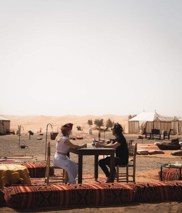 A man and a woman sit at an outside table at one of Desert Luxury Camp's sites in Morocco's Sahara Desert. The lady is dressed in white and is wearing a red headscarf, while the man is dressed in black with black sunglasses. There is a metallic teapot on their table. Colourful patterned cushions surround the table, and there are more seating areas partially visible in the near distance. Beyond that, we can see one of the site's luxury tents, and then the open desert beyond.